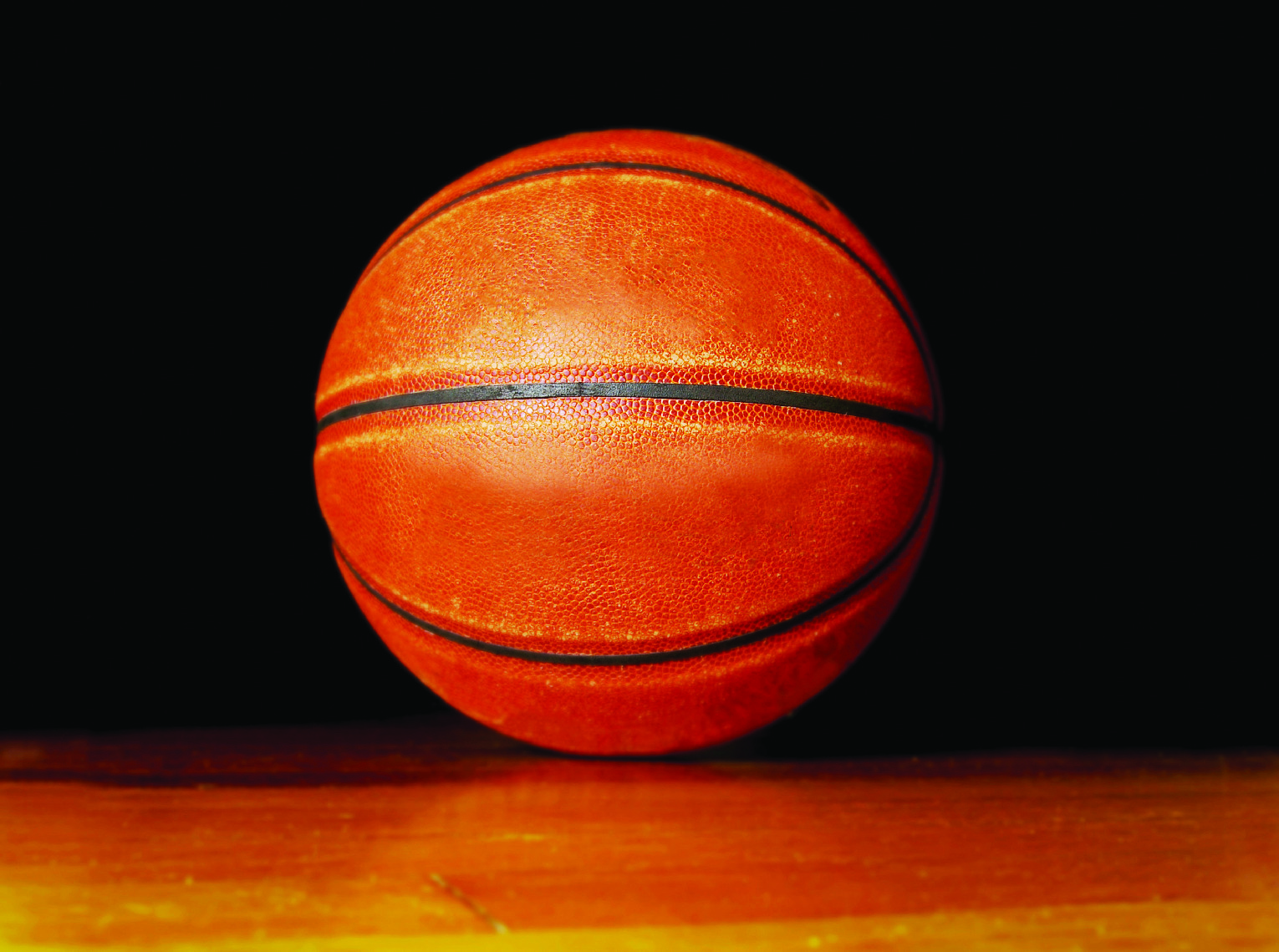 Close-up of an orange basketball resting on a wooden floor against a black background.