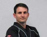 A studio-style portrait of a man wearing a black martial arts uniform with arms crossed, standing against a plain light-colored background. The uniform includes visible rank insignia on the sleeve, and the subject is facing the camera with a composed expression.