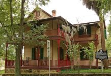 Walking through history at the Riley House The John G. Riley House, a historic two-story wood-frame home with wraparound porches, red railings, green shutters, and surrounding trees.