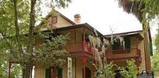 The John G. Riley House, a historic two-story wood-frame home with wraparound porches, red railings, green shutters, and surrounding trees.