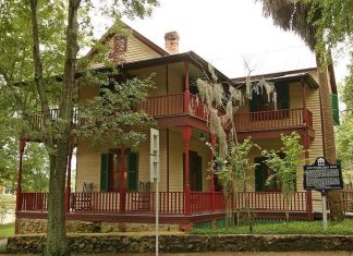 The John G. Riley House, a historic two-story wood-frame home with wraparound porches, red railings, green shutters, and surrounding trees.