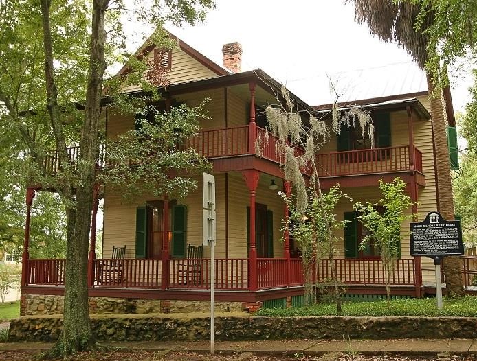 John G. Riley House The John G. Riley House, a historic two-story wood-frame home with wraparound porches, red railings, green shutters, and surrounding trees.