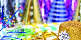 Gold glitter top hat with a “Happy New Year!” label sits on a table surrounded by colorful party hats, streamers, and confetti, suggesting a New Year’s celebration.