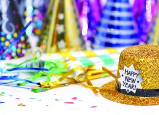 Gold glitter top hat with a “Happy New Year!” label sits on a table surrounded by colorful party hats, streamers, and confetti, suggesting a New Year’s celebration.