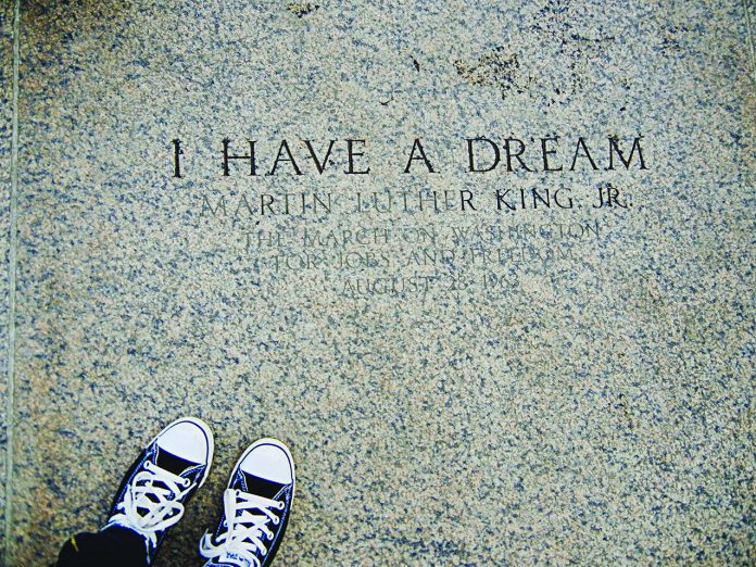 A photograph of engraved stone reading “I HAVE A DREAM – Martin Luther King Jr.”, referencing the March on Washington for Jobs and Freedom on August 28, 1963. The image is framed from above, with a pair of black-and-white sneakers visible at the bottom edge, creating a reflective, symbolic perspective on the historic inscription.