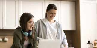 A woman and a teenage girl stand together in a bright kitchen, smiling as they look at a laptop on the counter. The teenager, wearing glasses and headphones around her neck, holds a pen and notepad, while the woman prepares food beside a bowl of salad ingredients.