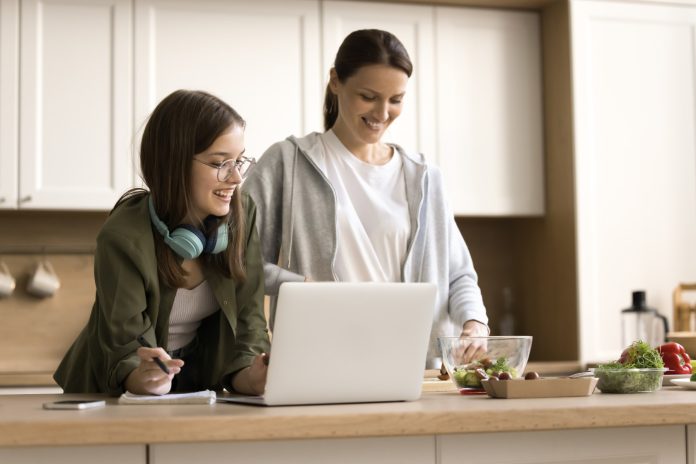 A woman and a teenage girl stand together in a bright kitchen, smiling as they look at a laptop on the counter. The teenager, wearing glasses and headphones around her neck, holds a pen and notepad, while the woman prepares food beside a bowl of salad ingredients.