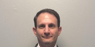 Professional waist-up portrait of a man with short dark hair standing against a plain gray background. He is wearing a dark gray suit jacket, a white button-down collared shirt, and a textured red tie.