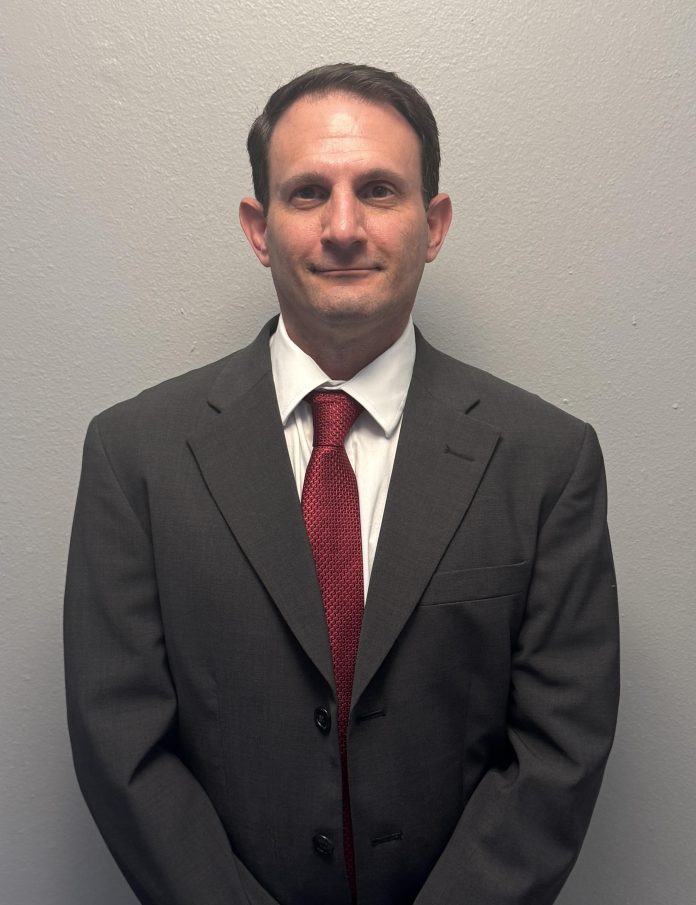 Professional waist-up portrait of a man with short dark hair standing against a plain gray background. He is wearing a dark gray suit jacket, a white button-down collared shirt, and a textured red tie.