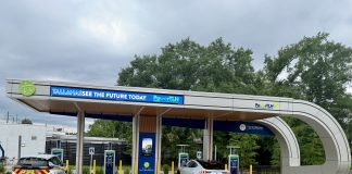 An outdoor electric vehicle charging station operated by the City of Tallahassee under a modern, curved canopy. A white Ford Mustang Mach-E and a white Chevrolet Bolt city utility vehicle are parked in charging bays. The canopy features digital signs displaying 'TALLAHASSEE SEE THE FUTURE TODAY' and 'PowerTLH' logos against a backdrop of trees and an overcast sky.