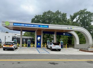 An outdoor electric vehicle charging station operated by the City of Tallahassee under a modern, curved canopy. A white Ford Mustang Mach-E and a white Chevrolet Bolt city utility vehicle are parked in charging bays. The canopy features digital signs displaying 'TALLAHASSEE SEE THE FUTURE TODAY' and 'PowerTLH' logos against a backdrop of trees and an overcast sky.