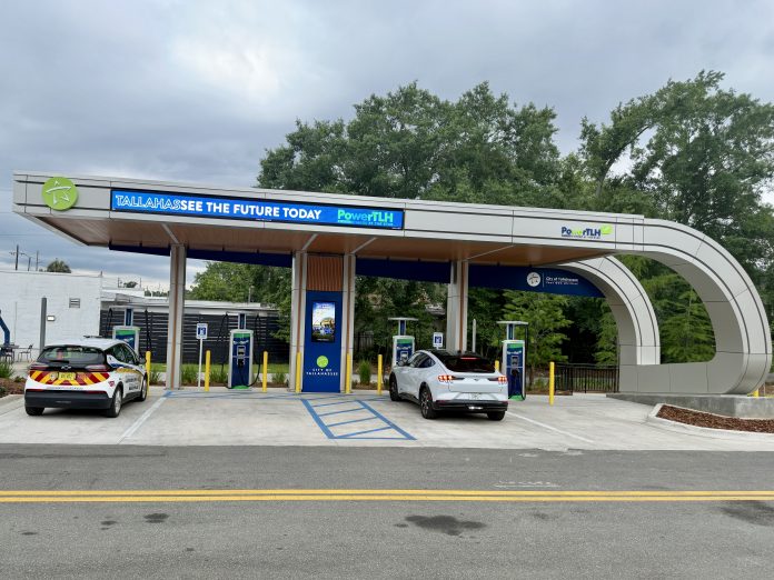 An outdoor electric vehicle charging station operated by the City of Tallahassee under a modern, curved canopy. A white Ford Mustang Mach-E and a white Chevrolet Bolt city utility vehicle are parked in charging bays. The canopy features digital signs displaying 'TALLAHASSEE SEE THE FUTURE TODAY' and 'PowerTLH' logos against a backdrop of trees and an overcast sky.