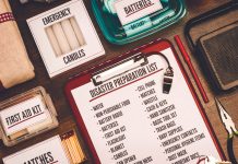 A flat-lay view of emergency supplies arranged on a wooden table around a red clipboard with a 'Disaster Preparation List.' The checklist includes items like water, non-perishable food, a battery radio, batteries, a first aid kit, and matches. Surrounding the clipboard are physical prep items including boxes labeled 'Emergency Candles,' 'First Aid Kit,' and 'Matches,' alongside a clear container of batteries, a radio, a whistle, a pen, and a pair of pliers.