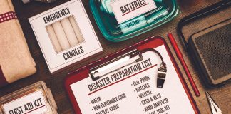 A flat-lay view of emergency supplies arranged on a wooden table around a red clipboard with a 'Disaster Preparation List.' The checklist includes items like water, non-perishable food, a battery radio, batteries, a first aid kit, and matches. Surrounding the clipboard are physical prep items including boxes labeled 'Emergency Candles,' 'First Aid Kit,' and 'Matches,' alongside a clear container of batteries, a radio, a whistle, a pen, and a pair of pliers.