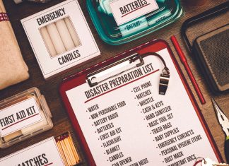 A flat-lay view of emergency supplies arranged on a wooden table around a red clipboard with a 'Disaster Preparation List.' The checklist includes items like water, non-perishable food, a battery radio, batteries, a first aid kit, and matches. Surrounding the clipboard are physical prep items including boxes labeled 'Emergency Candles,' 'First Aid Kit,' and 'Matches,' alongside a clear container of batteries, a radio, a whistle, a pen, and a pair of pliers.