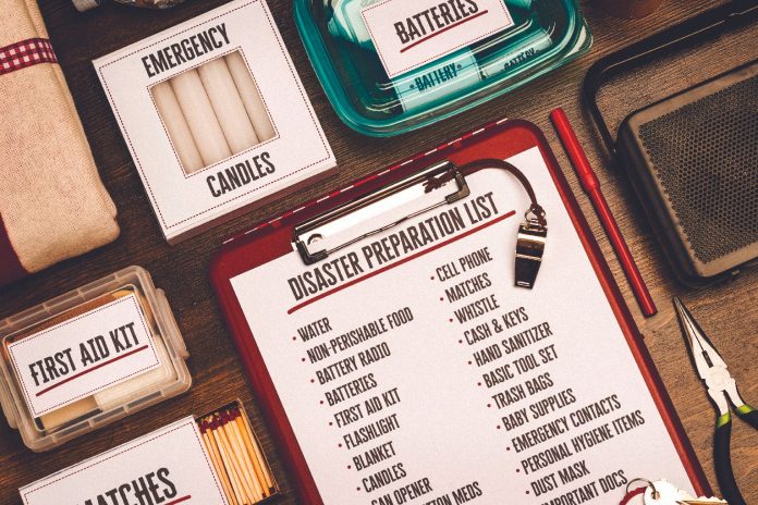 N2412P62002C A flat-lay view of emergency supplies arranged on a wooden table around a red clipboard with a 'Disaster Preparation List.' The checklist includes items like water, non-perishable food, a battery radio, batteries, a first aid kit, and matches. Surrounding the clipboard are physical prep items including boxes labeled 'Emergency Candles,' 'First Aid Kit,' and 'Matches,' alongside a clear container of batteries, a radio, a whistle, a pen, and a pair of pliers.