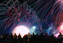 A vibrant nighttime photograph showing a silhouette of a crowd of spectators looking up at a massive, colorful fireworks display. Bright pink, blue, and green light trails arc across the black sky above a brilliant white burst of light at the center.