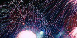 A vibrant nighttime photograph showing a silhouette of a crowd of spectators looking up at a massive, colorful fireworks display. Bright pink, blue, and green light trails arc across the black sky above a brilliant white burst of light at the center.