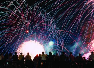 A vibrant nighttime photograph showing a silhouette of a crowd of spectators looking up at a massive, colorful fireworks display. Bright pink, blue, and green light trails arc across the black sky above a brilliant white burst of light at the center.