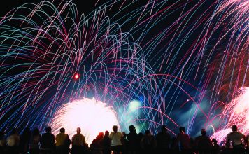 A vibrant nighttime photograph showing a silhouette of a crowd of spectators looking up at a massive, colorful fireworks display. Bright pink, blue, and green light trails arc across the black sky above a brilliant white burst of light at the center.
