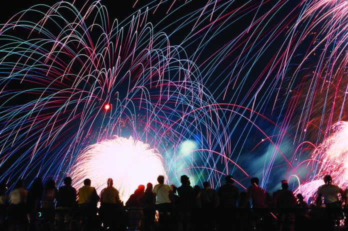 A vibrant nighttime photograph showing a silhouette of a crowd of spectators looking up at a massive, colorful fireworks display. Bright pink, blue, and green light trails arc across the black sky above a brilliant white burst of light at the center.