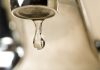 Annual report confirms Tallahassee drinking water meets all quality standards An extreme close-up photograph of a single, clear droplet of water hanging from the aerator tip of a chrome faucet just before falling. The background is softly blurred with warm, neutral tones.