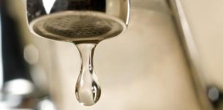 An extreme close-up photograph of a single, clear droplet of water hanging from the aerator tip of a chrome faucet just before falling. The background is softly blurred with warm, neutral tones.