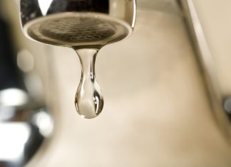 An extreme close-up photograph of a single, clear droplet of water hanging from the aerator tip of a chrome faucet just before falling. The background is softly blurred with warm, neutral tones.