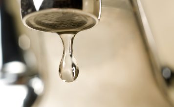 An extreme close-up photograph of a single, clear droplet of water hanging from the aerator tip of a chrome faucet just before falling. The background is softly blurred with warm, neutral tones.